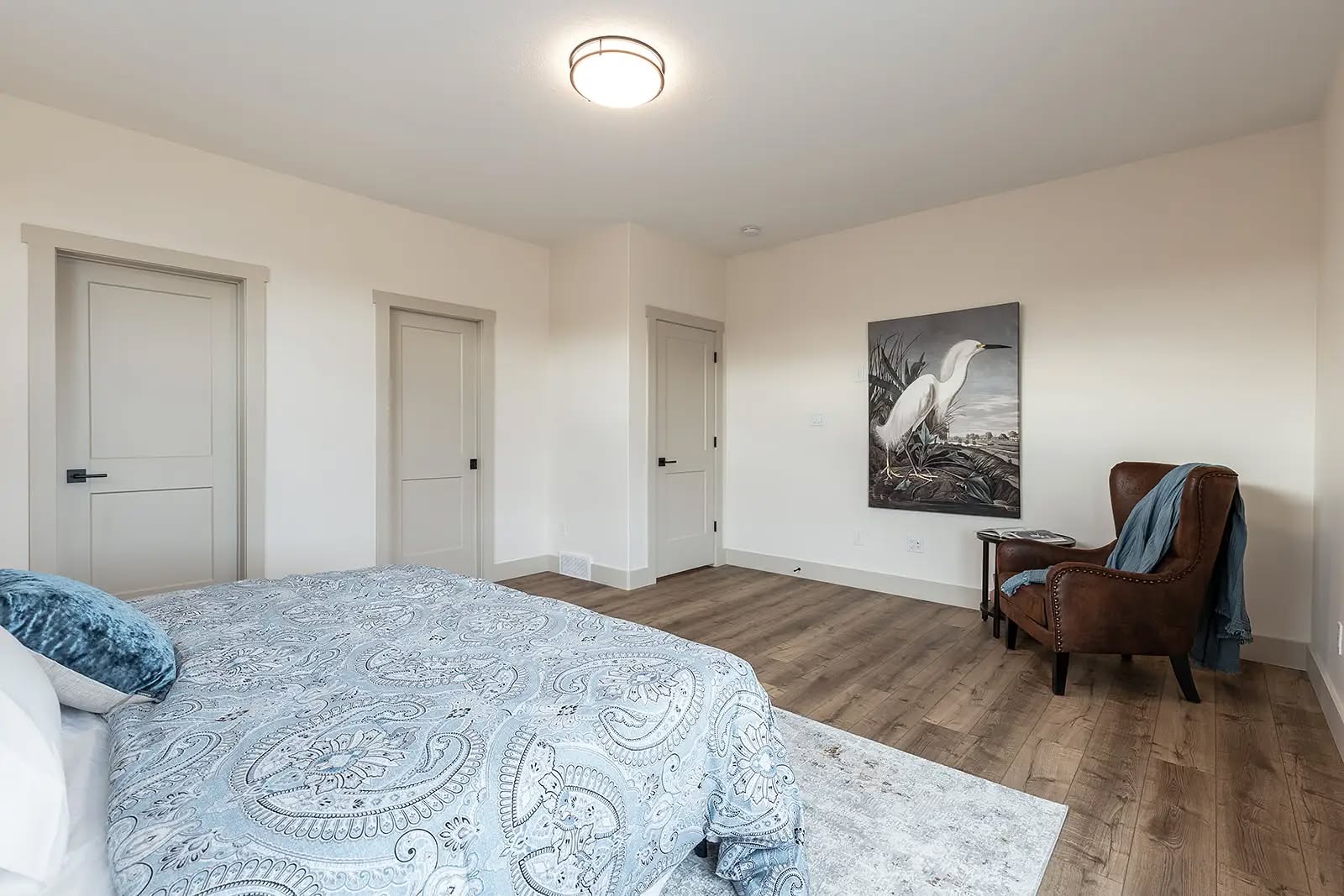 A cozy bedroom with a patterned blue bedspread, wooden floor, and a brown armchair with a blue blanket. A heron painting adds a peaceful touch.