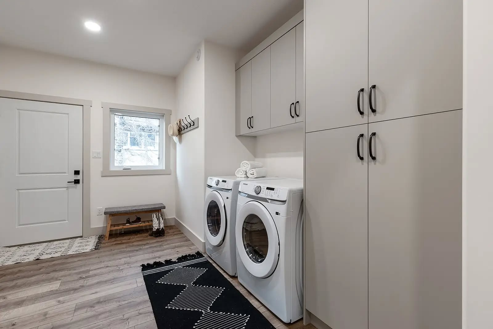 Modern laundry room with white washer and dryer, overhead and tall storage cabinets, a black patterned rug, wooden bench, and window with hooks.