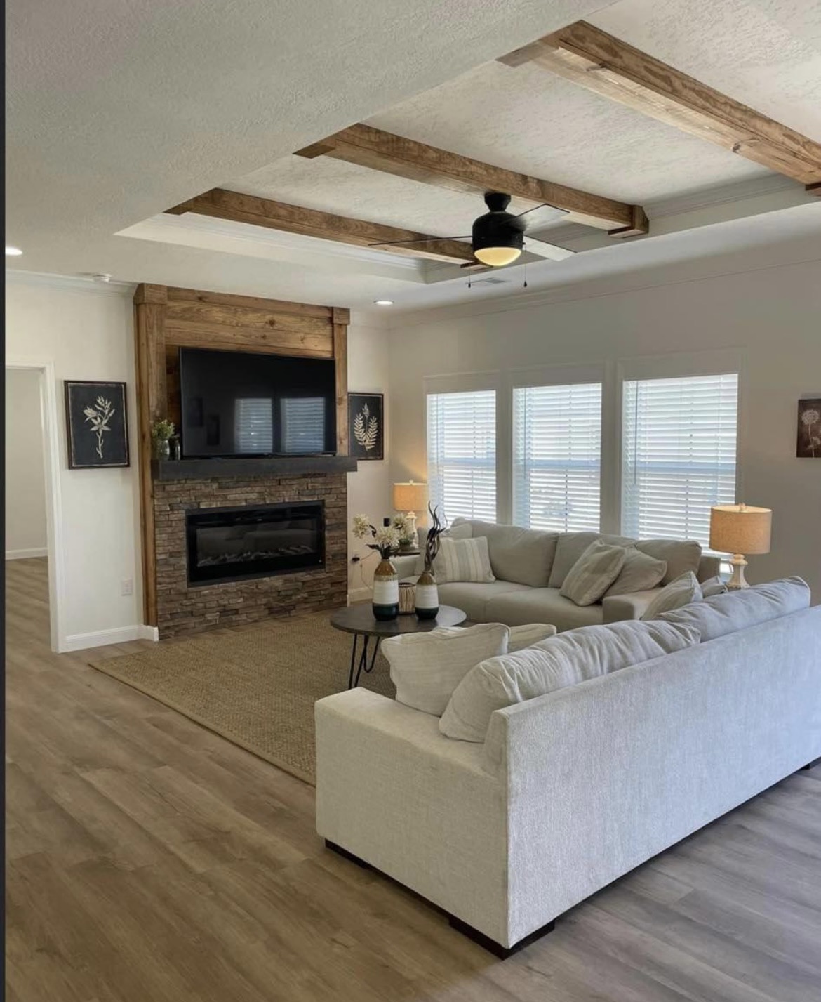Modern living room with light wood floors, beige sectional sofa, and stone fireplace. Ceiling has wooden beams and fan, creating a cozy atmosphere.