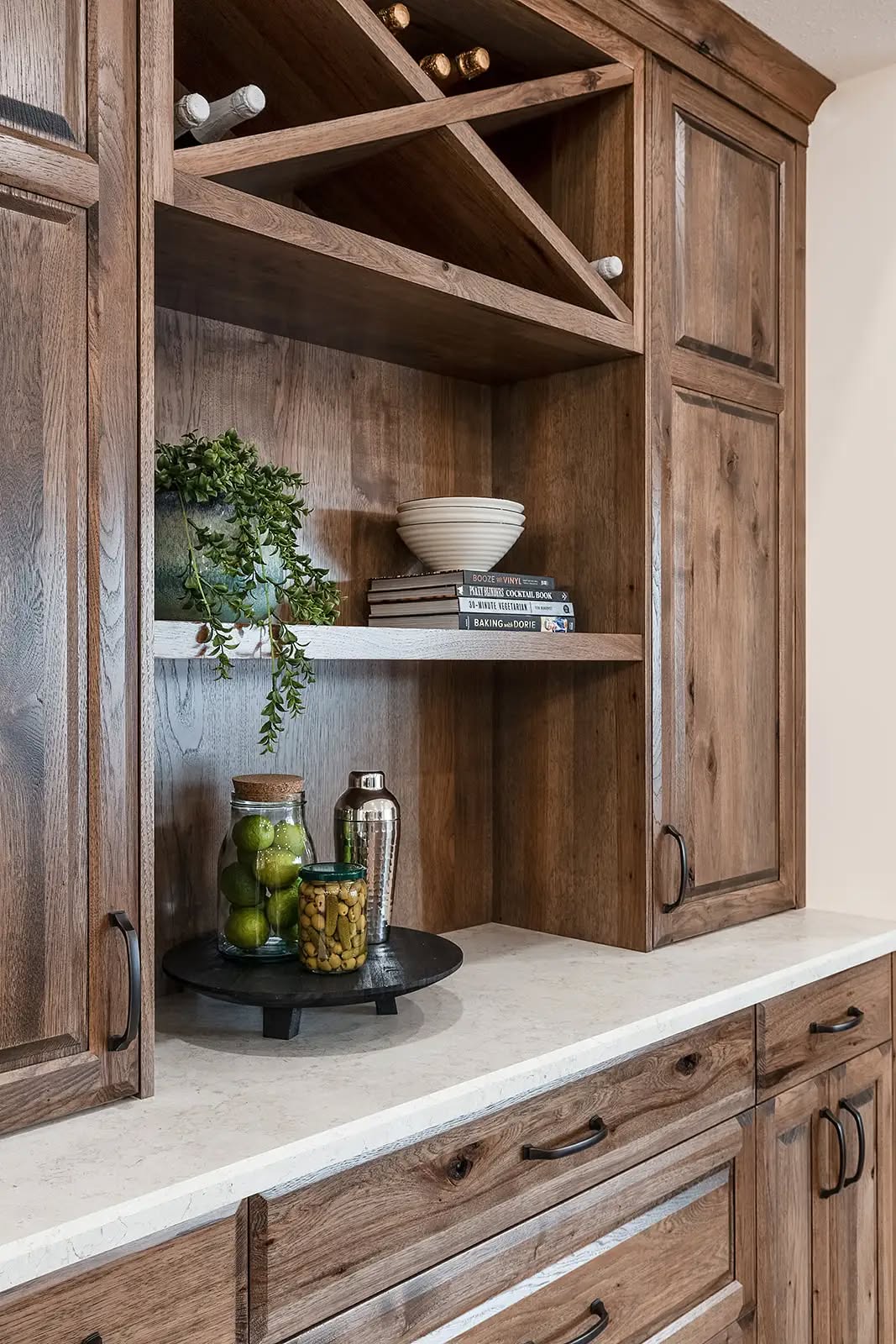 Rustic kitchen cabinet with natural wood finish, open shelves holding a white bowl, books, greenery, a tray with jars and a cocktail shaker. Cozy ambiance.