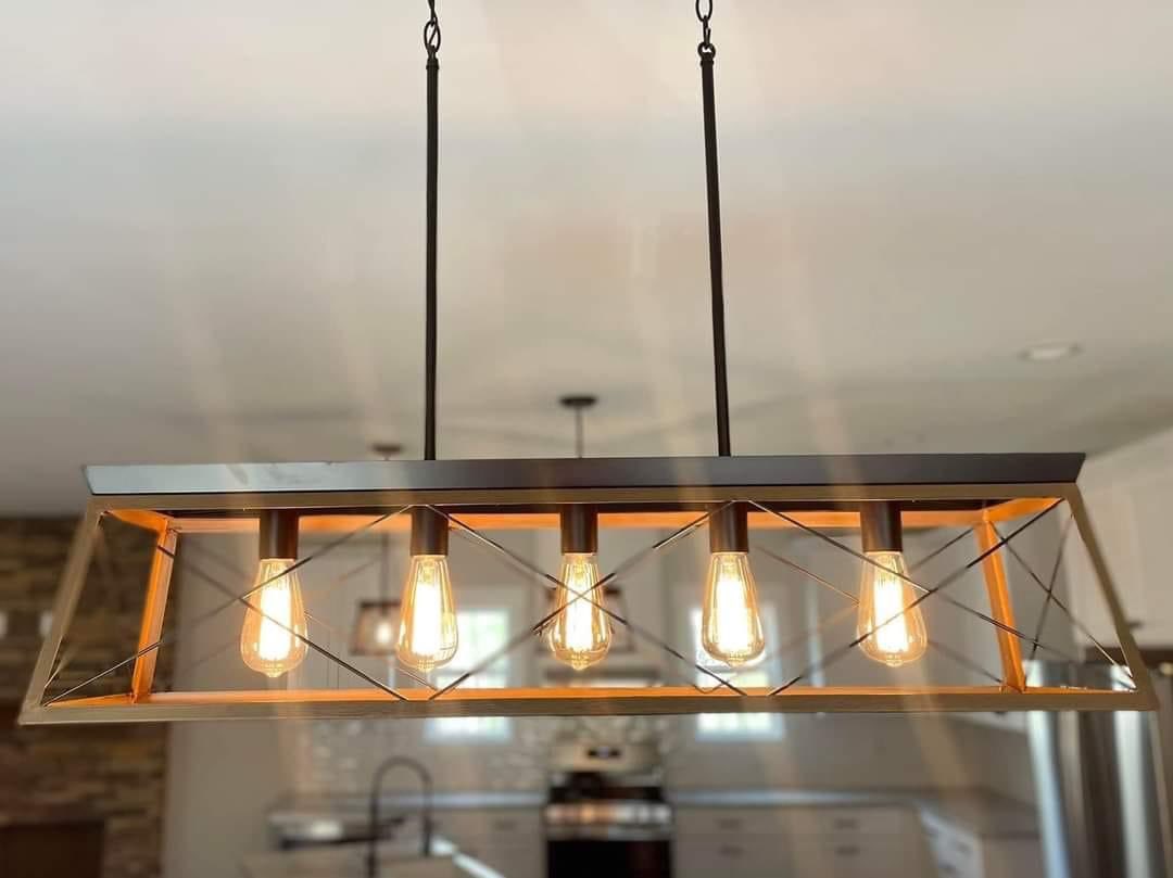 A rectangular pendant light fixture with five glowing Edison bulbs hangs from a ceiling. The modern design features metal rods in a kitchen setting.