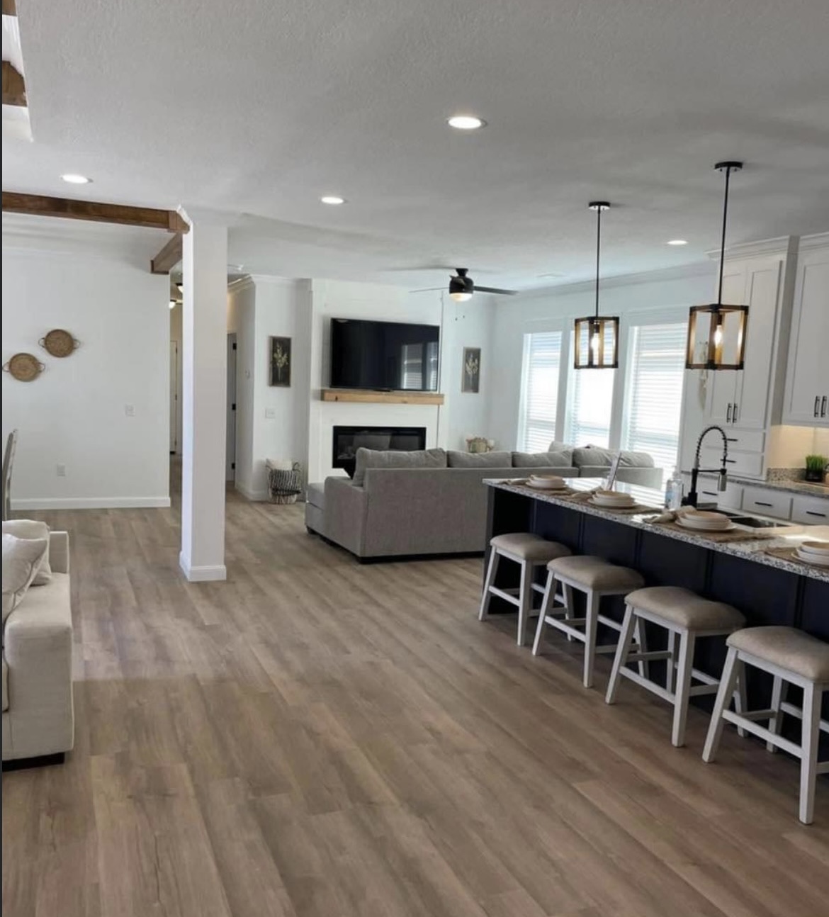 Spacious open-plan living room and kitchen with light wood floors, gray sofas, a large TV, and a kitchen island with stools, creating a cozy ambiance.