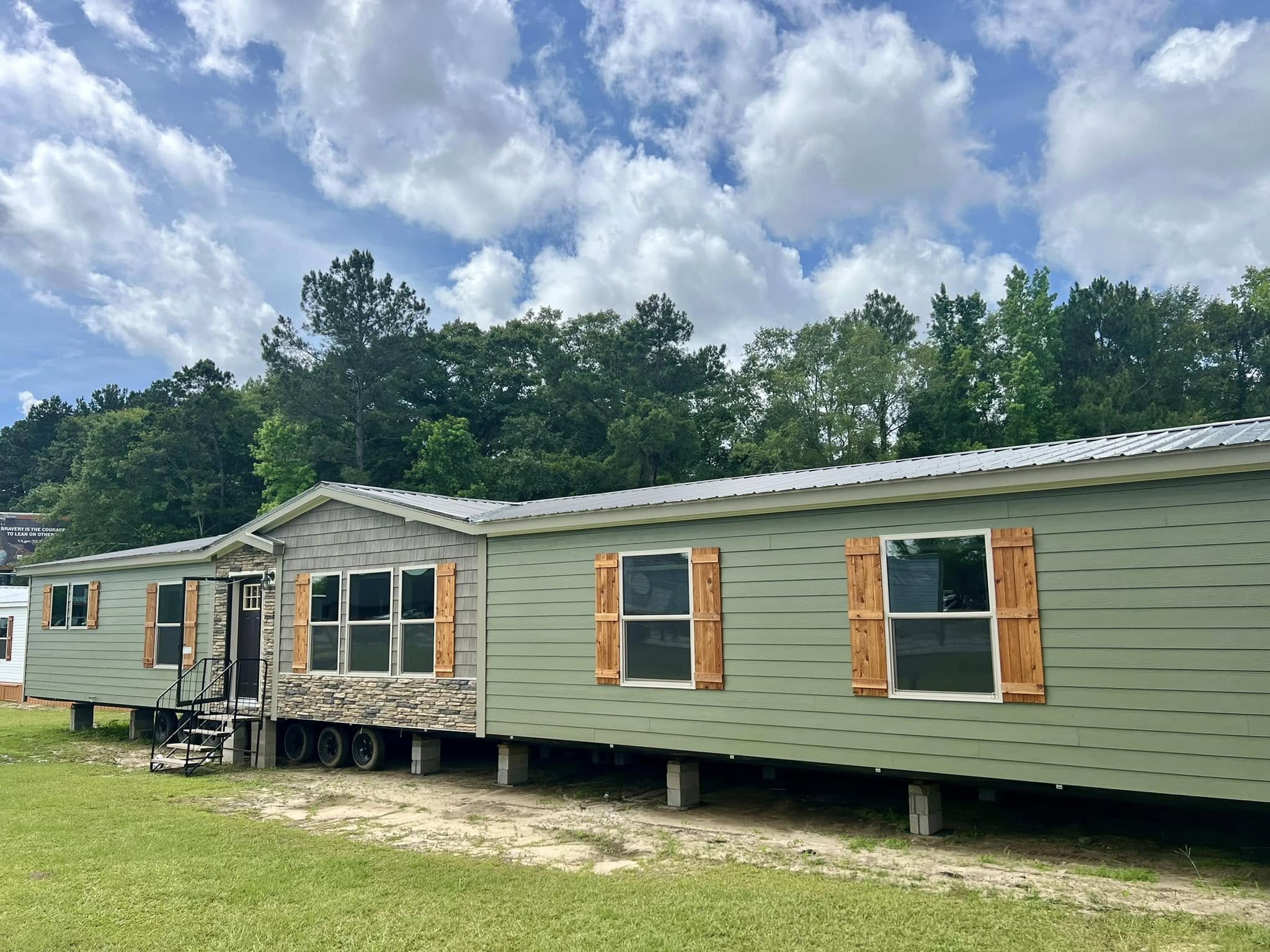 A large light green manufactured home with wooden shutters and a stone facade stands on a grassy lawn. Background features lush trees under a partly cloudy sky.