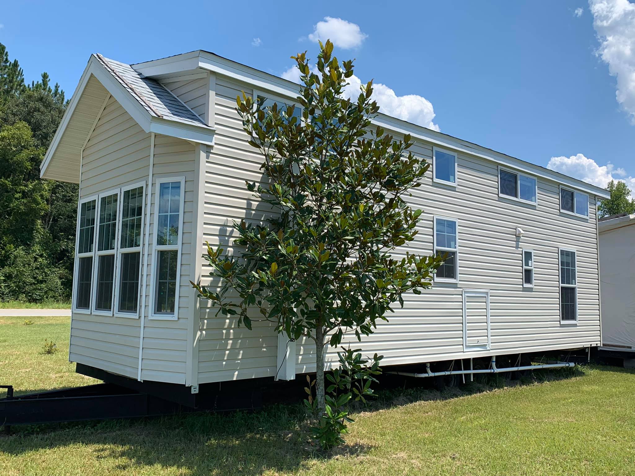 A beige mobile home with large front windows and a gabled roof is set against a clear blue sky. A small tree grows in the foreground, enhancing the serene, rural setting.