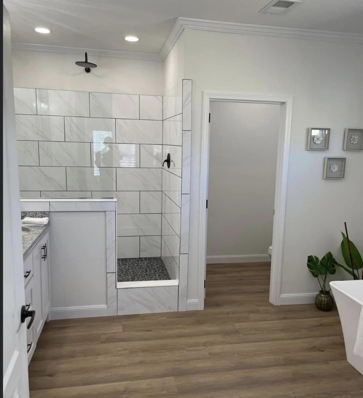 Modern bathroom with a wooden floor, white tiled shower featuring black fixtures, a white vanity with granite countertop, and a small potted plant.