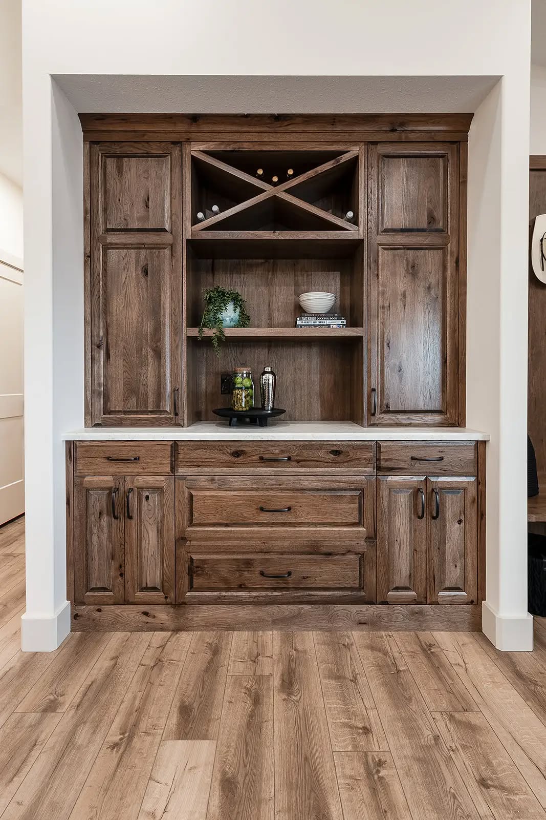Rustic wooden cabinet set against a beige wall, featuring a wine rack, shelves with books and plants, and drawers. Warm tones convey a cozy, inviting ambiance.