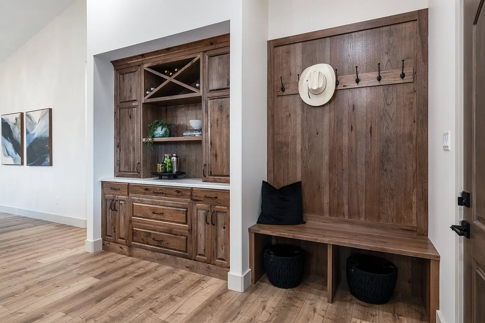 Rustic entryway with wood cabinetry and a bench. Shelves display decor, and hooks hold a white hat. Light wood flooring adds warmth.
