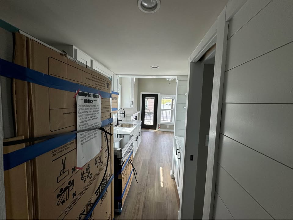 Narrow kitchen under renovation with unpacked appliances lining the left side and white cabinets on the right. Bright light through the glass door.