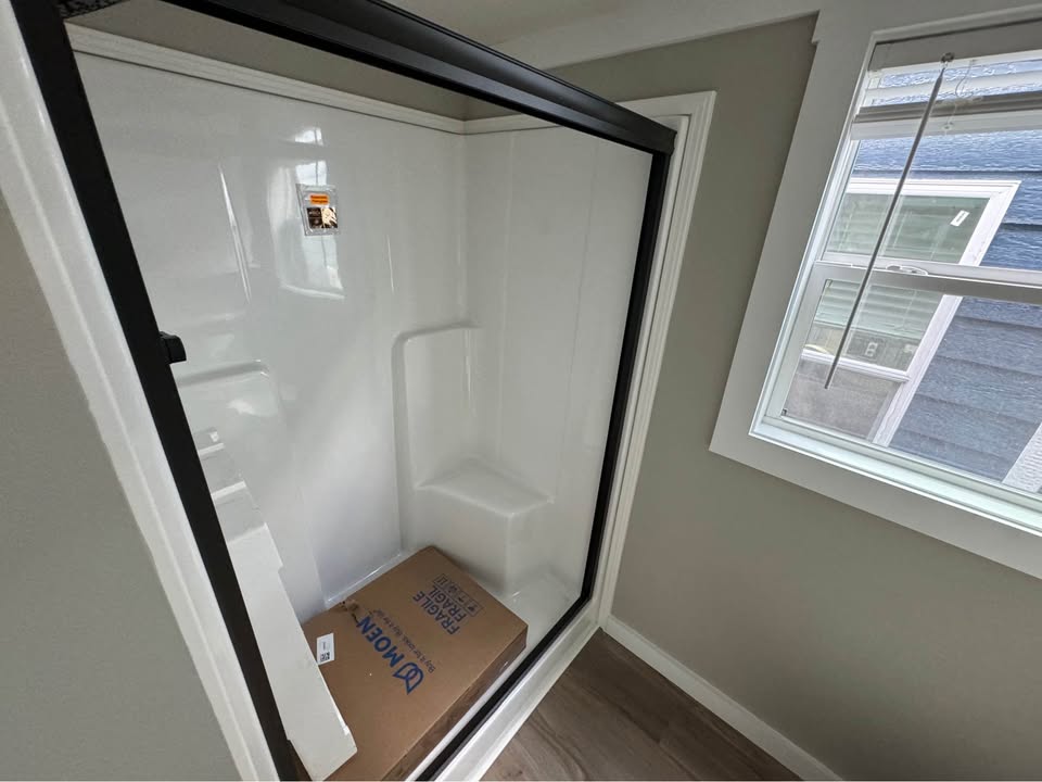 A newly installed white shower with a glass door and black frame, featuring a built-in seat and a cardboard box inside. Natural light comes from a nearby window.
