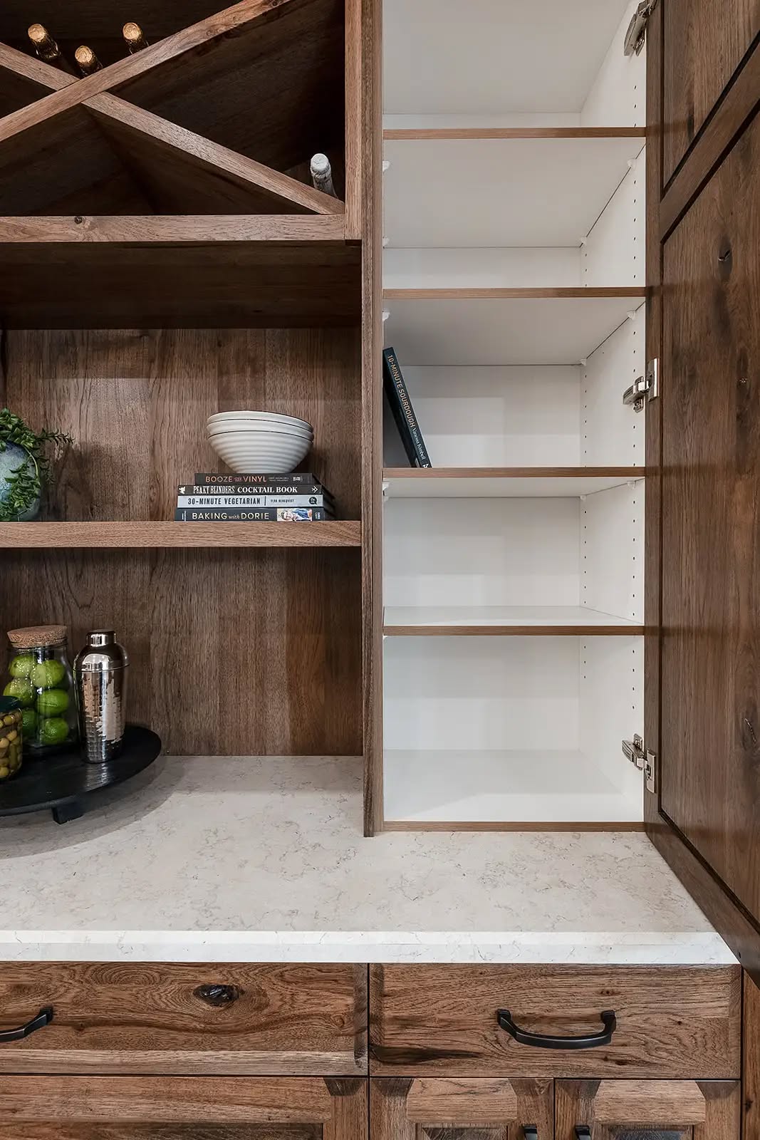 Rustic kitchen with wooden cabinets, wine rack, books, and a white bowl. Shelves hold a cocktail set and fresh limes. Open cupboard is empty. Cozy vibe.