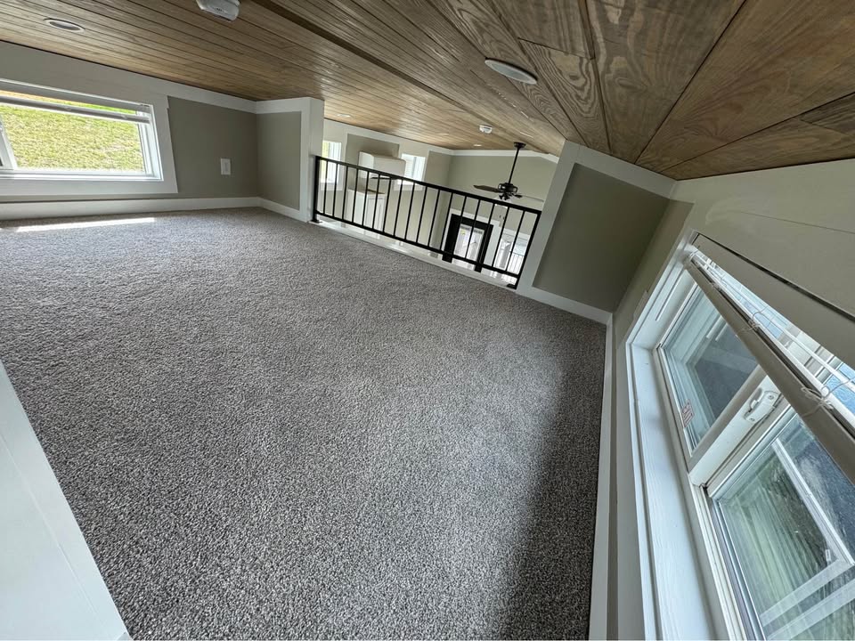 Cozy loft space with carpeted floor, wooden ceiling, and natural light from large windows. Black railing overlooks open living area below.