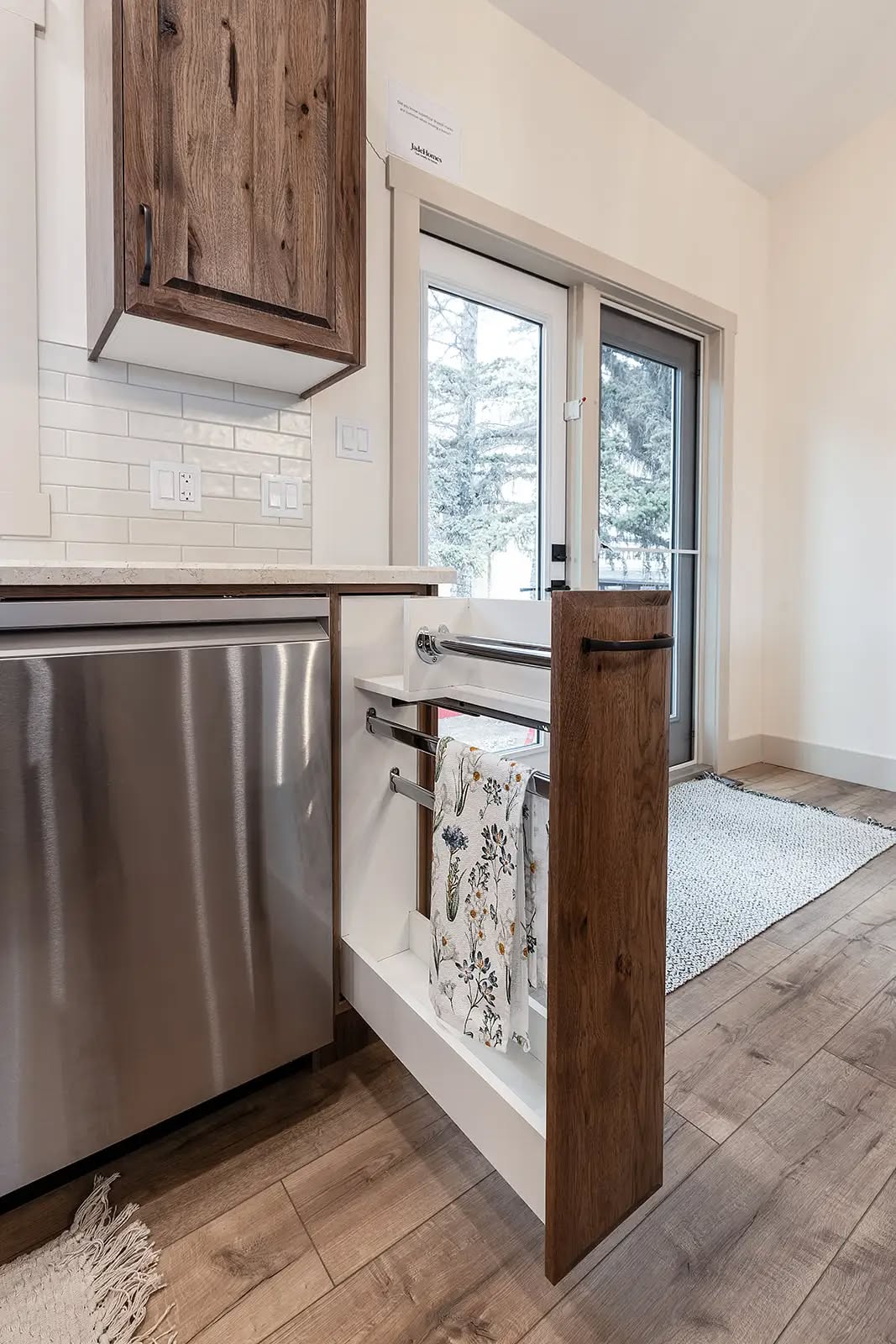 A modern kitchen with light wood floors and a sliding patio door. A pull-out cabinet reveals towel racks with floral-patterned towels, next to a dishwasher.