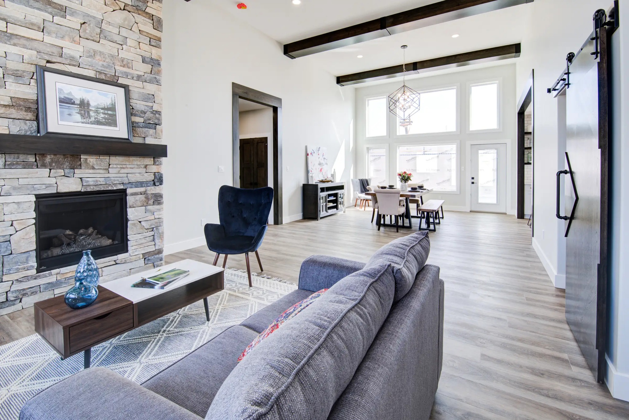 Modern open-concept living room with stone fireplace, gray sofa, and wooden coffee table. Dining area in background with large windows, exuding a bright, airy feel.