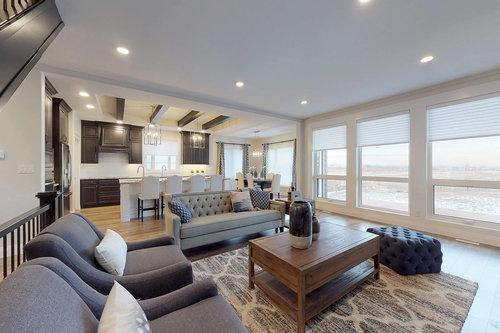 Spacious modern living room with large windows, gray sofas, and a wooden coffee table. Open kitchen with dark cabinets and bar stools in view. Cozy and bright.