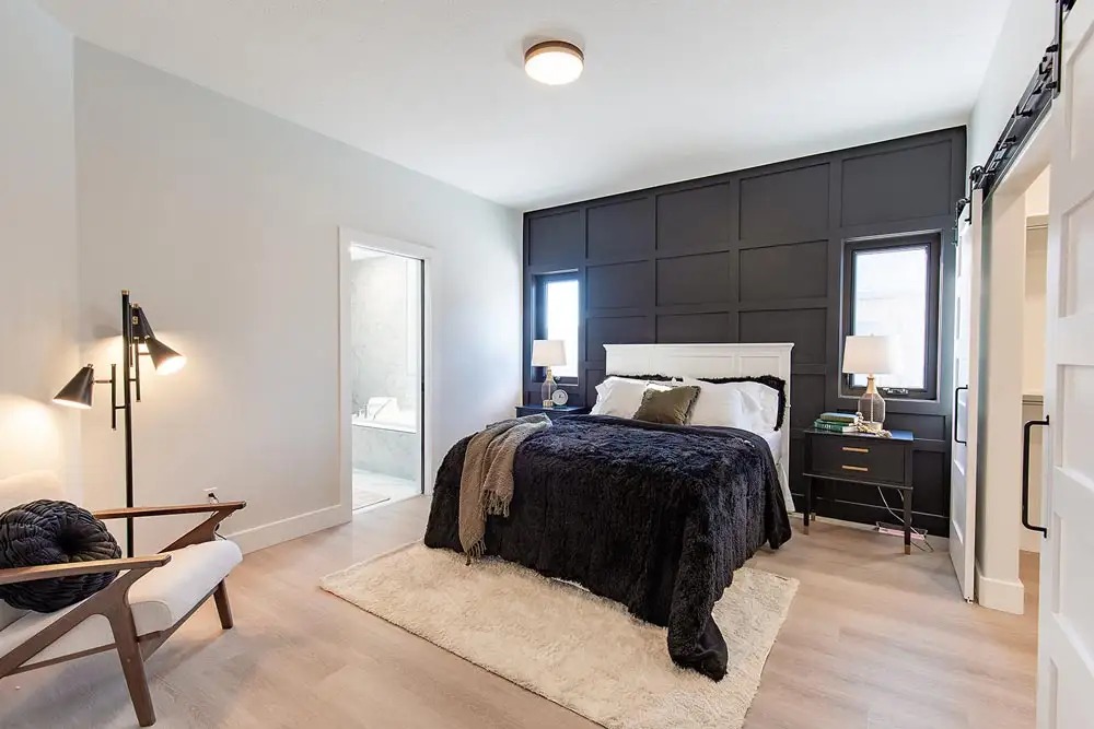 Modern bedroom with a cozy feel, featuring a dark paneled accent wall, plush black bedding, and warm lighting. A chair and floor lamp add stylish touches.