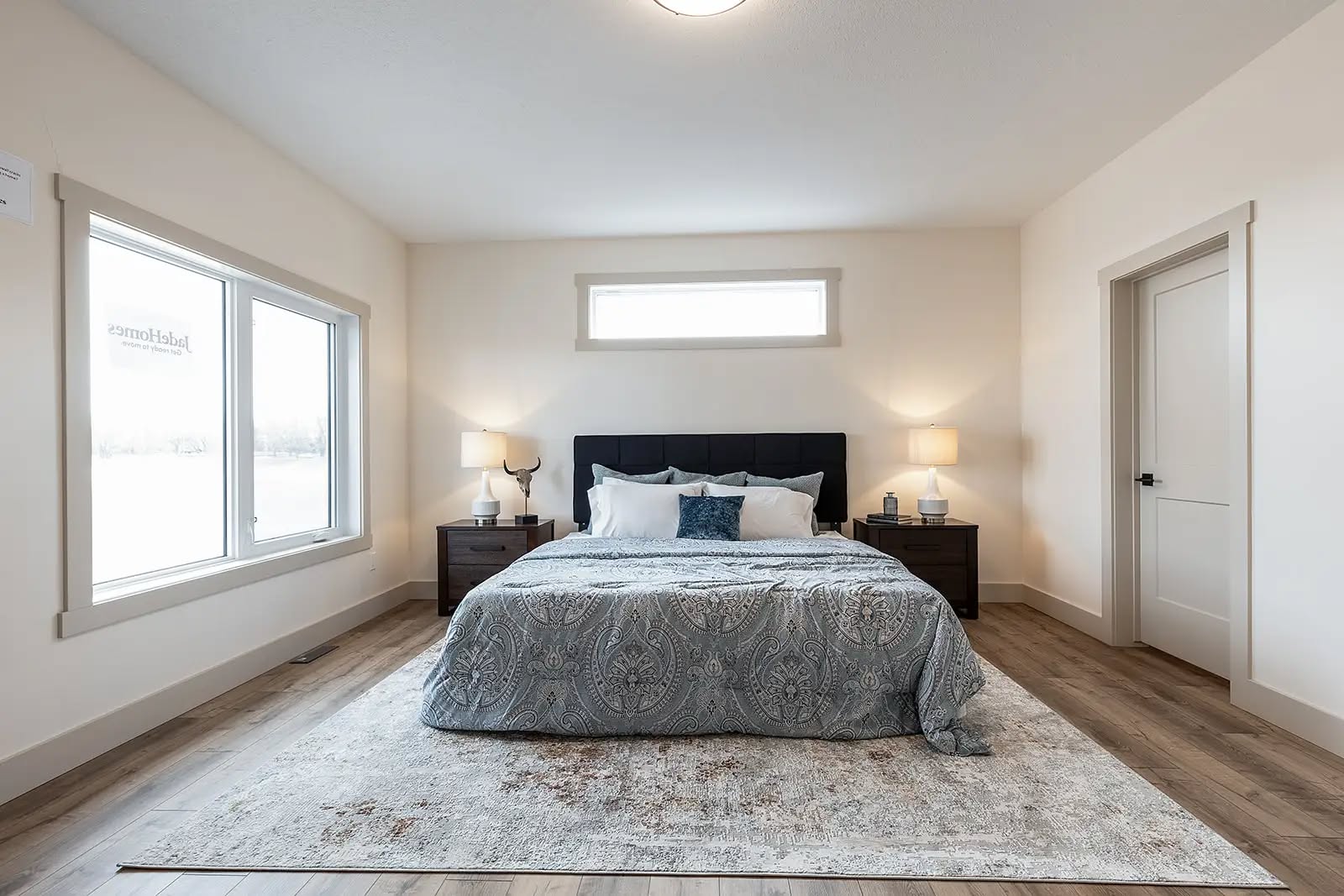 Modern bedroom with a cozy, patterned bedspread, flanked by wooden nightstands with lamps. Large windows and soft lighting create a serene atmosphere.