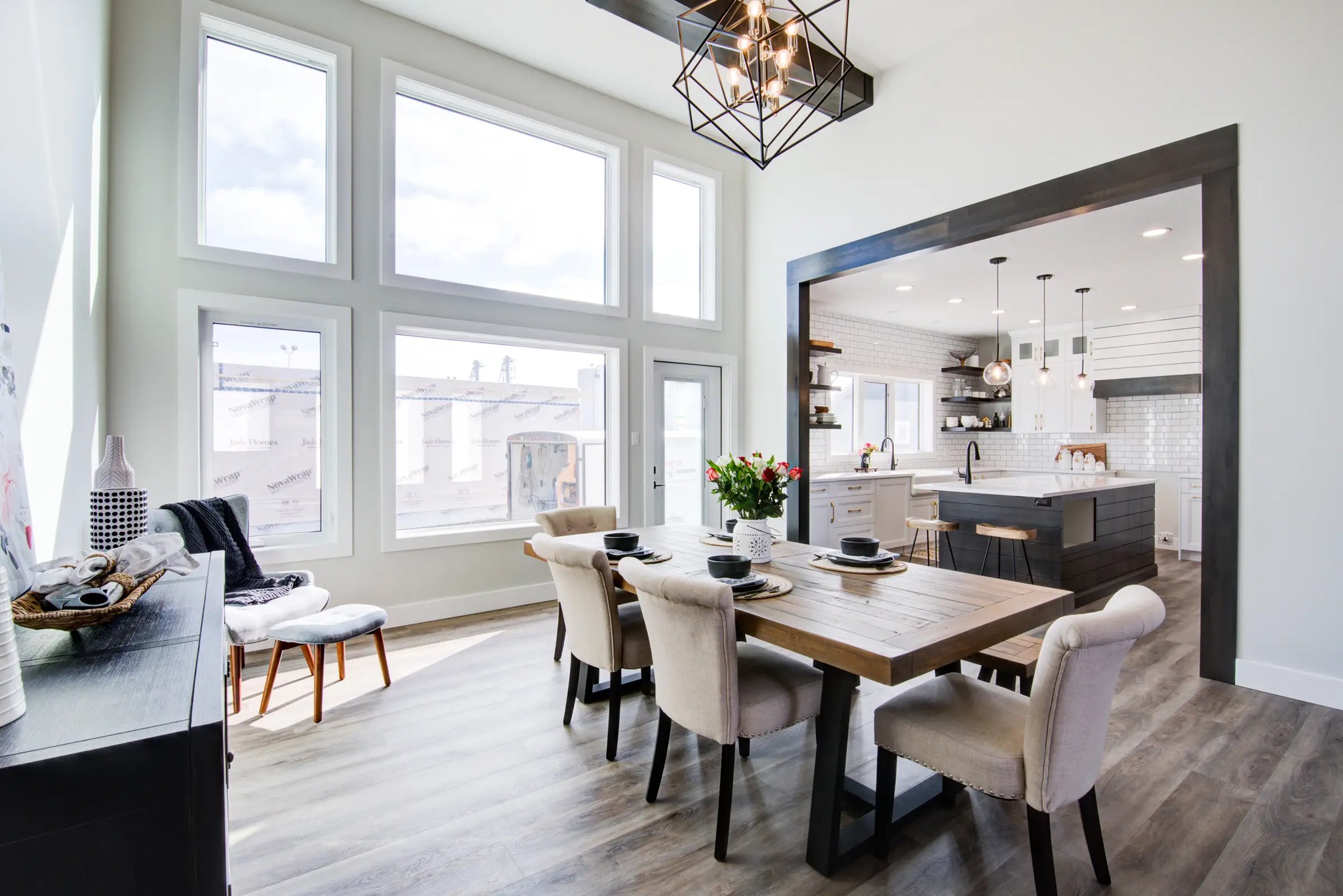 Bright dining area with a wooden table, six cushioned chairs, and a vase of tulips. Modern kitchen visible through a wide doorway, featuring pendant lights.