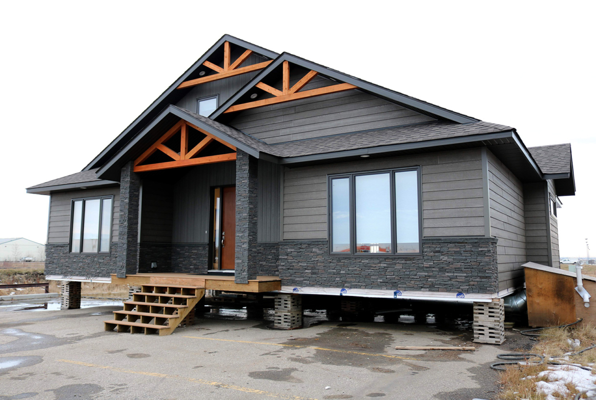A gray modular home elevated on blocks with a dark stone facade and wood accents. Wooden stairs lead to a front door, set against a cloudy sky.