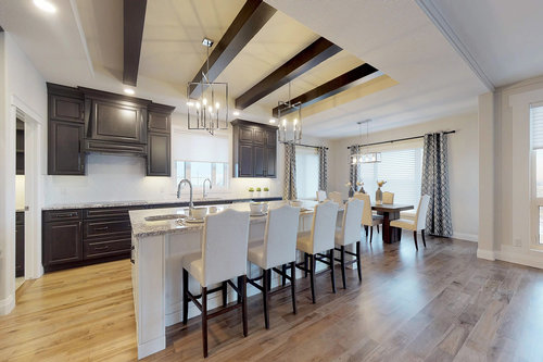 Modern kitchen and dining area with dark wood cabinets, white island, and bar stools. Large windows, hardwood floors, and pendant lighting create a bright, elegant atmosphere.