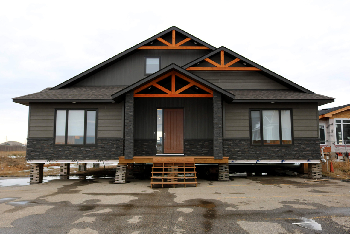 A modern single-story house with dark gray siding, wooden accents, and a central wooden door. Elevated on blocks, it has large windows and a triangular roof.