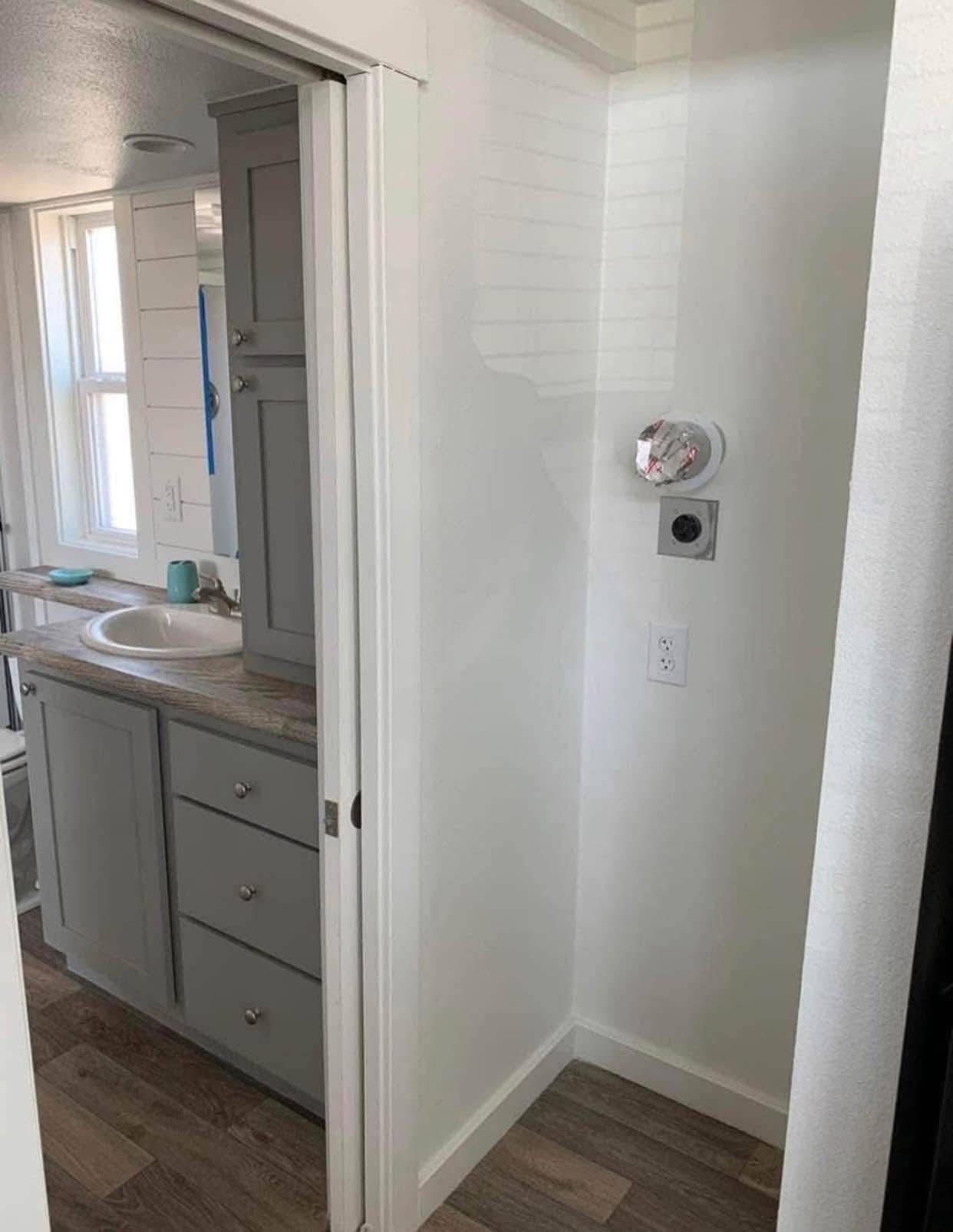 A narrow bathroom with gray cabinets and wood flooring. A partially open sliding door reveals a sink area. A smoke detector is awkwardly placed on a wall next to an electrical socket, adding a touch of humor.