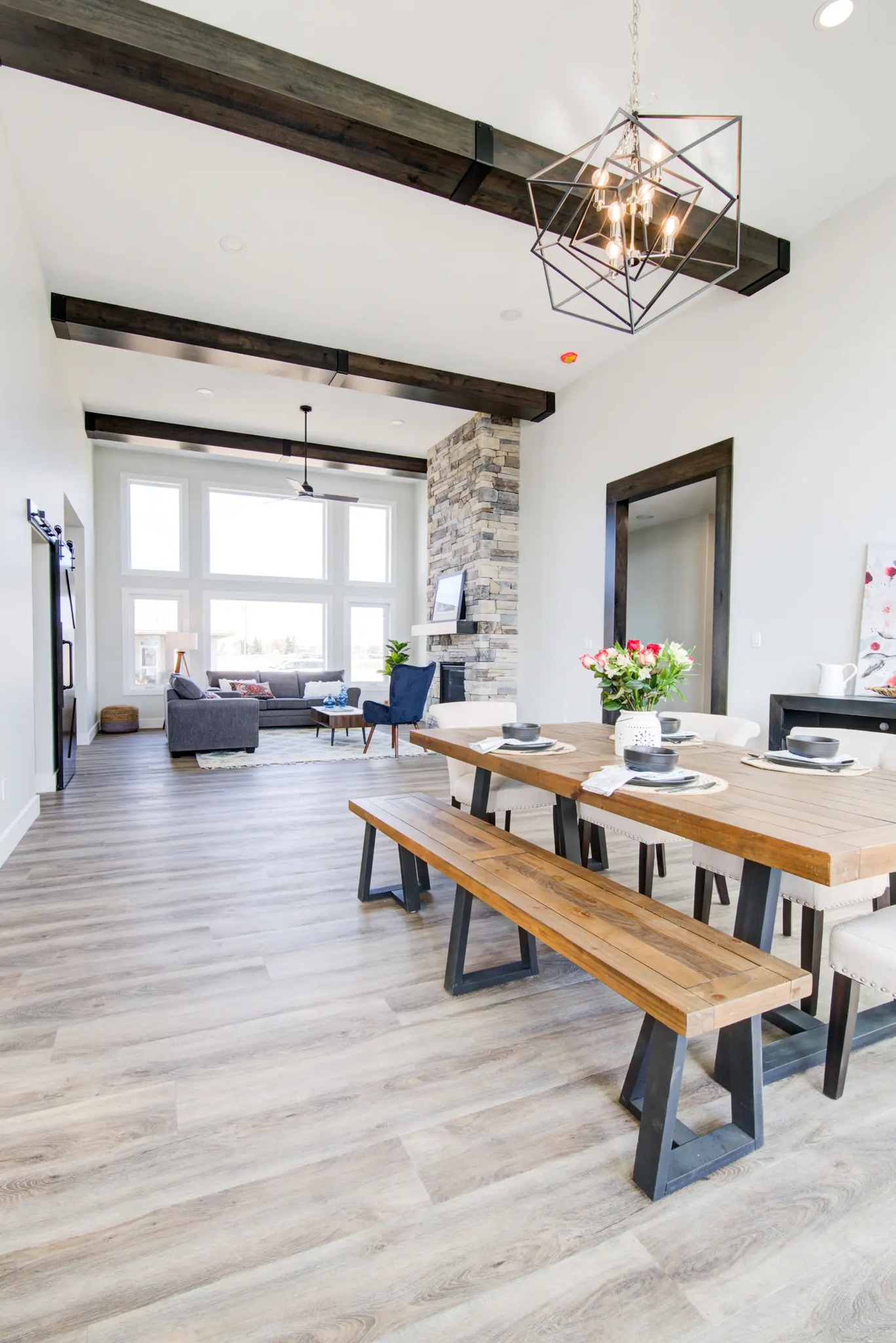 Modern open-concept living room and dining area with wooden flooring. Features a wooden dining table with benches, high ceiling, and large windows. Airy and bright.