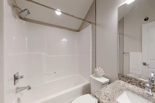 A modern bathroom features a white bathtub with a showerhead above. There's a granite countertop with a sink, mirror, and decorative coral. The ambiance is clean and minimalistic.