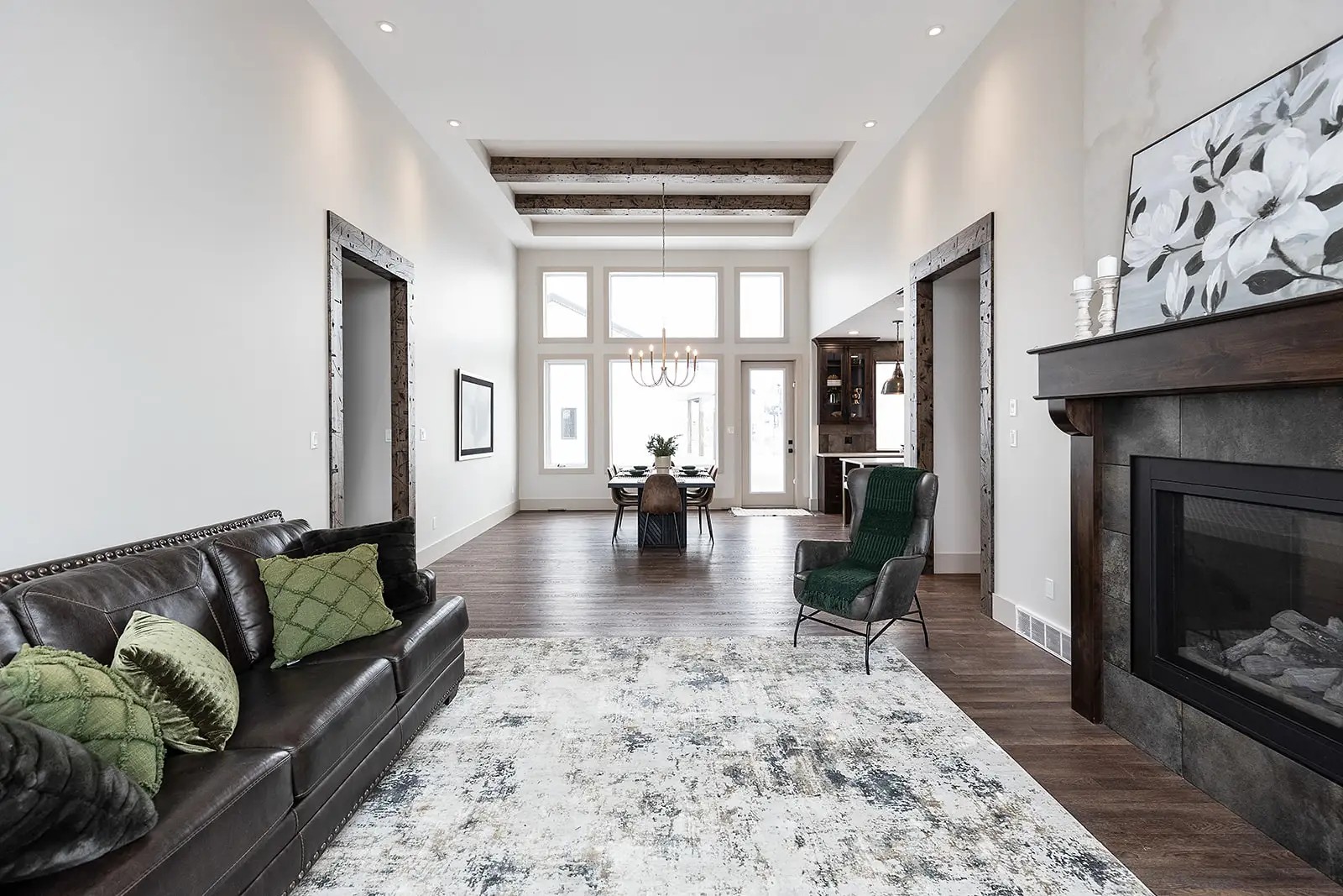 Modern living room with high ceilings, wooden beams, and large windows. Features a brown leather sofa with green pillows, a patterned rug, and a fireplace with floral artwork. The dining area has a wooden table and a chandelier, creating an elegant, cozy atmosphere.