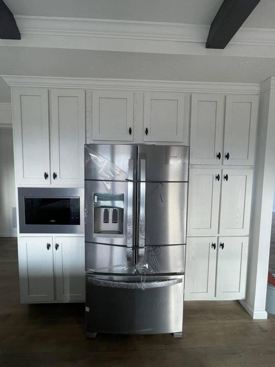 Stainless steel refrigerator wrapped in protective plastic stands in front of white paneled cabinets. Adjacent is a built-in microwave. The space is modern and minimalistic.