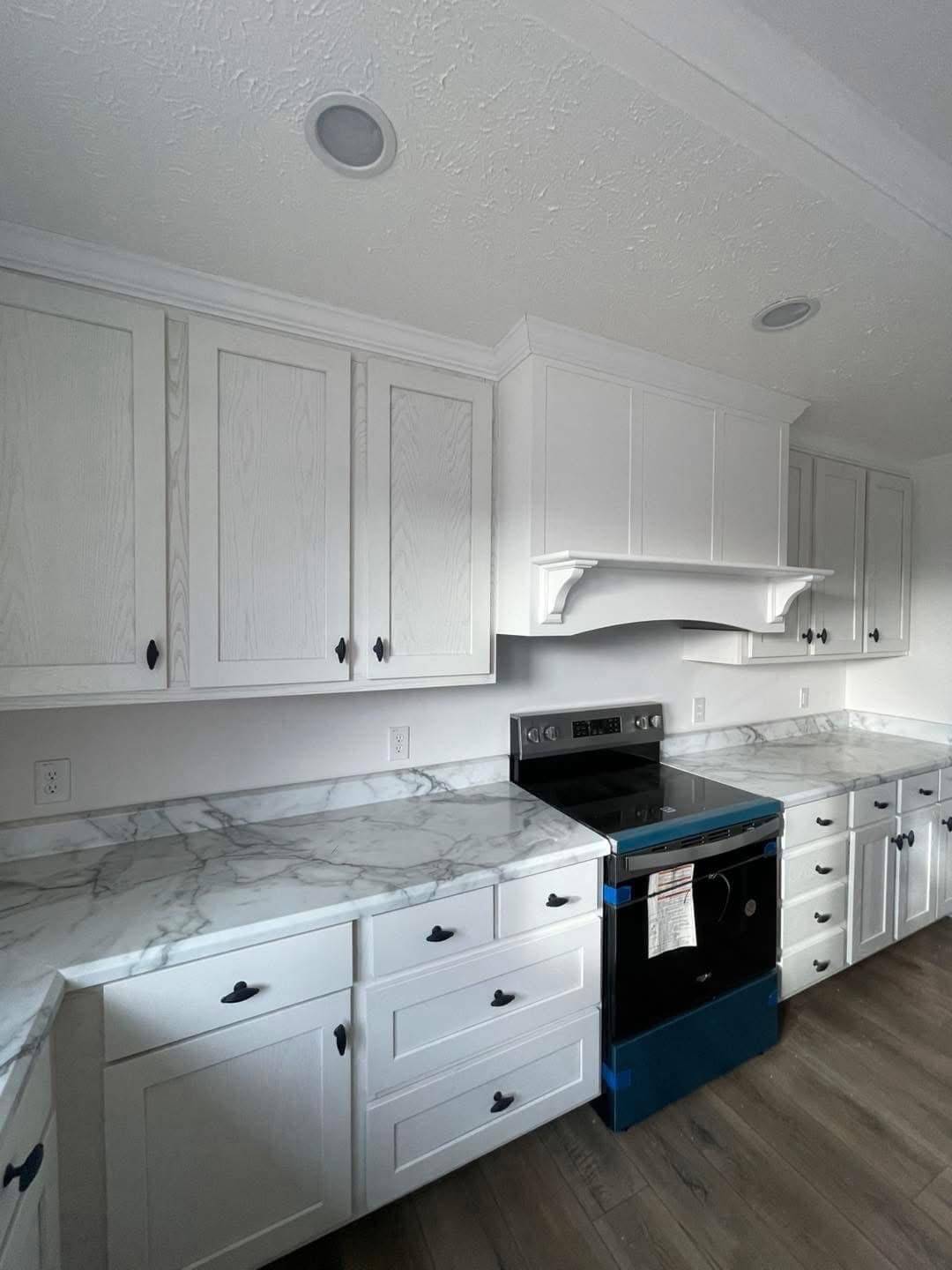 Modern kitchen with white cabinets and marble countertops. A new stainless steel oven is wrapped in blue protective film. The space feels clean and bright.