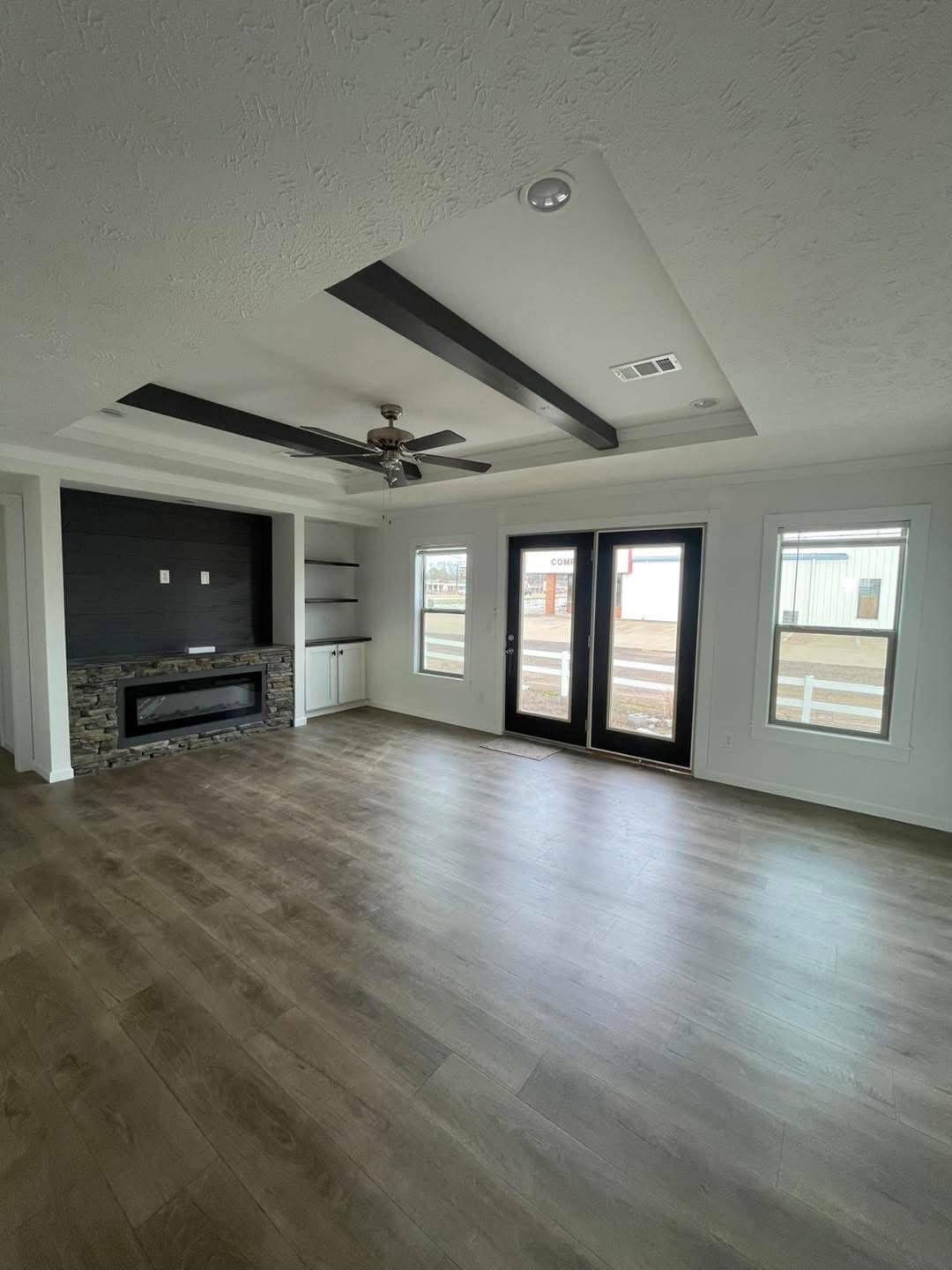 Spacious empty living room with wooden floors and a faux stone fireplace. Features a modern ceiling fan, recessed shelves, and large windows. Bright and airy.