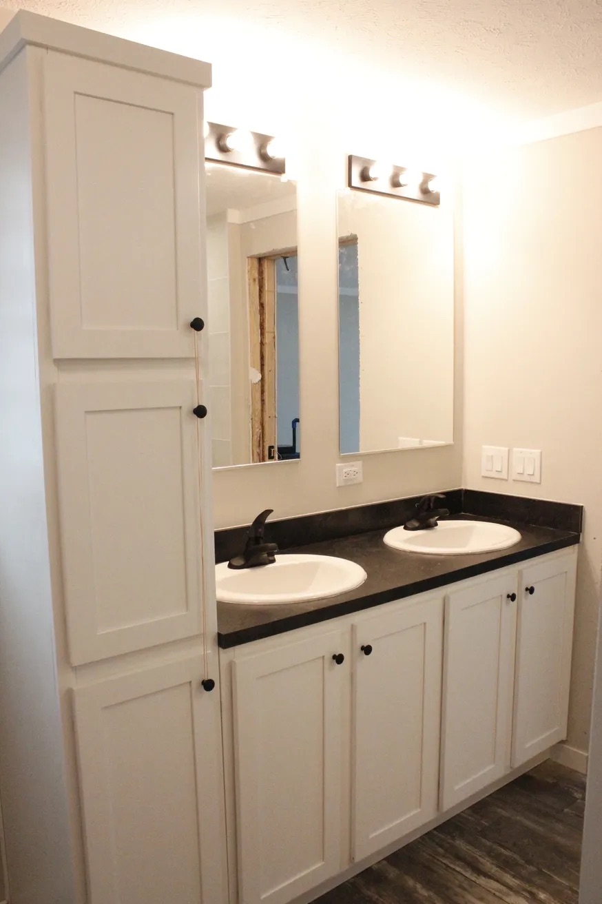 A modern bathroom with two white sinks on a black countertop. Above are two mirrors under bright lights. White cabinets flank each side, accentuating a clean, minimalistic design.