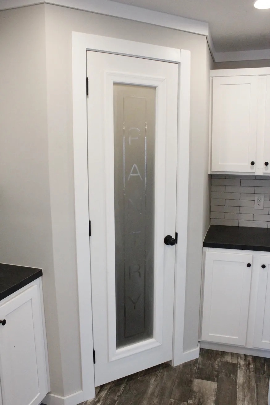 White kitchen pantry door with frosted glass displaying "PANTRY" vertically. Modern cabinets and dark countertops create a clean, organized look.