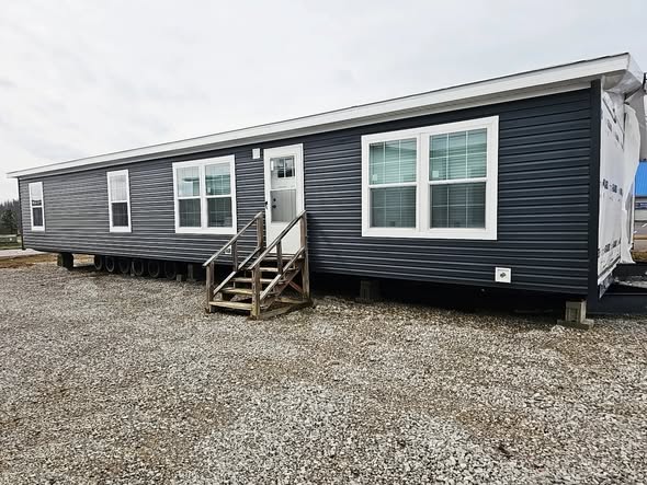 Long, dark gray modular home with white trim and multiple windows. Situated on gravel, with wooden steps leading to the door. Overcast sky.