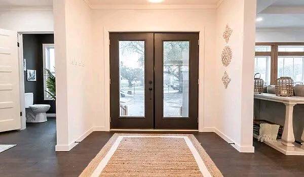 Bright and inviting entryway with double glass doors framed by dark wood. A woven rug leads to the doors. Adjacent rooms feature modern decor.