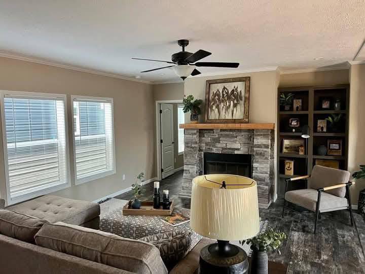 Cozy living room with a gray sectional, stone fireplace, and a ceiling fan. Shelves with decor, a beige armchair, and two large windows create an inviting atmosphere.