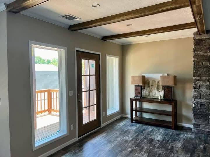 Bright room with wooden ceiling beams, a glass door, and two vertical windows leading to a deck. Stone feature wall and wooden console table with lamps.