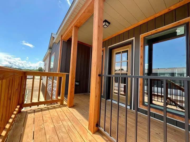 Wooden deck with railing on a sunny day, featuring a dark cabin exterior with large windows and a glass door leading inside. The mood is warm and inviting.