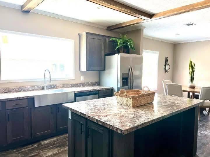 A modern kitchen with dark wood cabinets and marble countertops. A large island with a wicker basket is centered. Stainless steel appliances and a dining area with beige chairs are in the background. A window lets in natural light, highlighting a clean and inviting space.