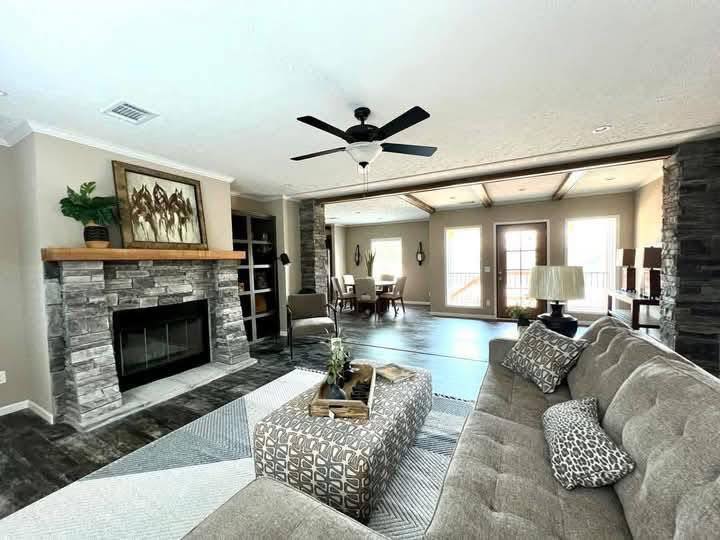 Spacious living room with stone fireplace, wooden mantel, and ceiling fan. A beige sectional sofa and patterned ottoman create a cozy, inviting atmosphere.