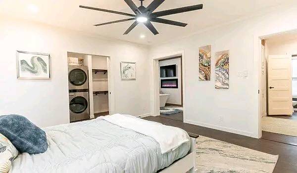 A modern bedroom with a white bed and gray throw, ceiling fan, and art on white walls. Open closet with stacked washer and dryer. Calm, minimalist tone.