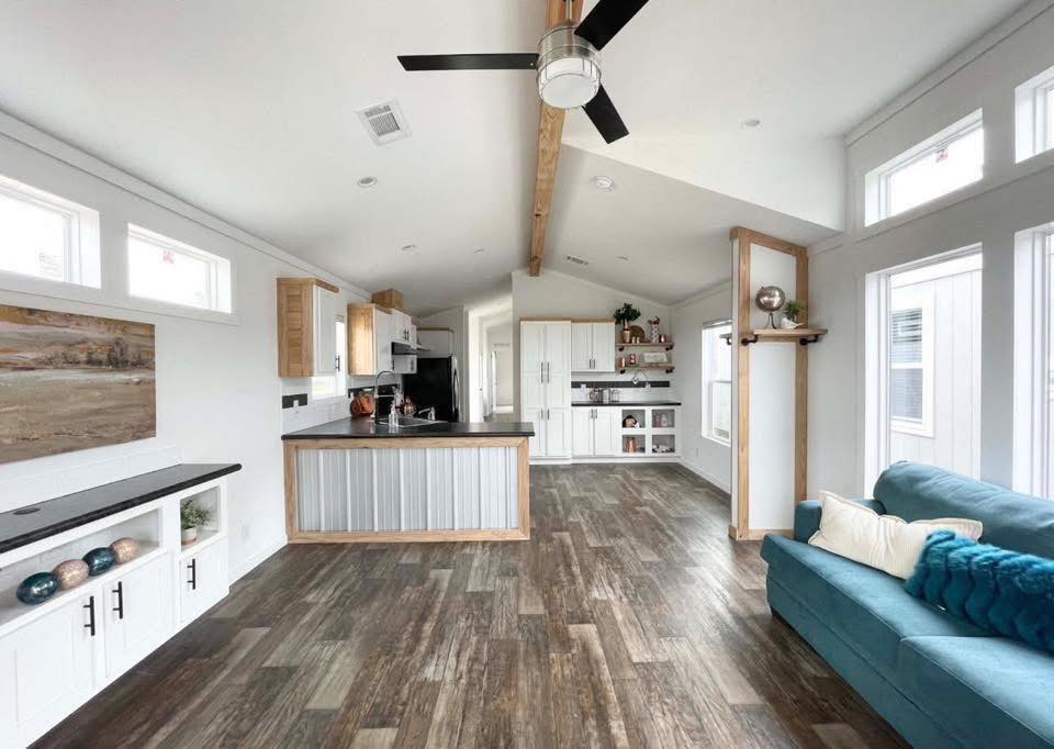 Spacious living area with high ceiling, wood accents, and ceiling fan. Features rustic kitchen, wood floors, teal sofa with pillows, and abundant light.