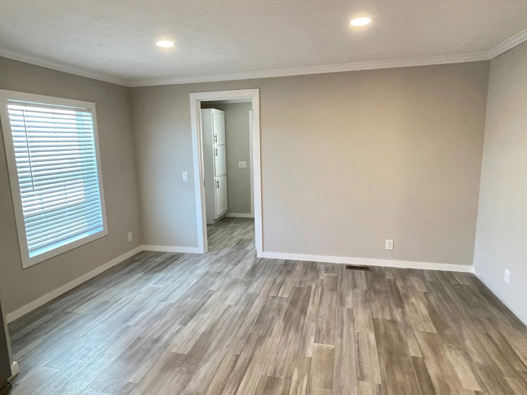 A bright, empty room with beige walls and wood-look flooring. A large window with blinds lets in natural light. An open doorway leads to a kitchen.