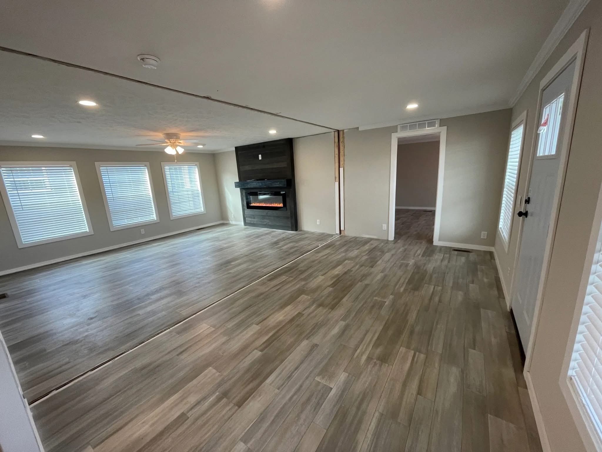 Spacious room with light wood flooring, white walls, and a black fireplace on one side. Three large windows and a ceiling fan provide brightness.