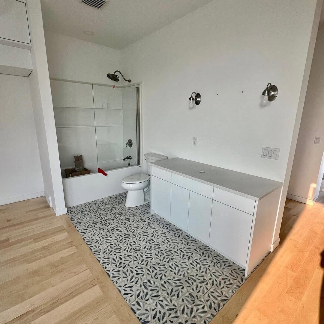 Bright bathroom with patterned tile flooring, a bathtub, glass shower screen, white vanity, and wall-mounted lights. Minimalist and modern design.