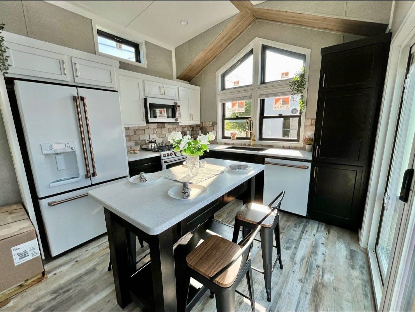 Modern kitchen with high ceilings and large windows. Features a center island with stools, white appliances, wood accents, and potted greenery, creating a bright, cozy atmosphere.