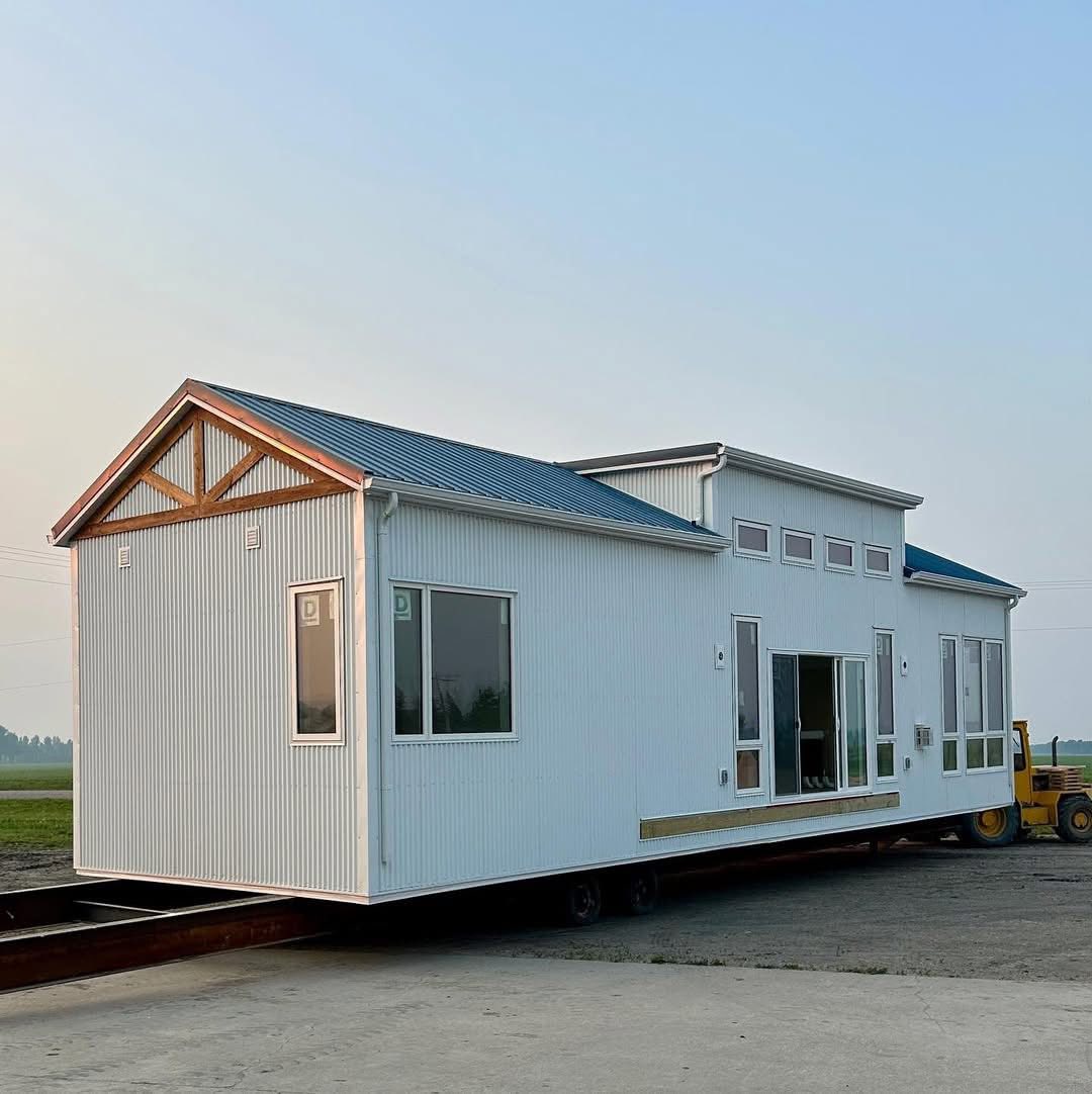 A white tiny house with a gabled roof is on a trailer in an outdoor setting. Large windows and a sliding door accentuate its modern design. The sky is clear.