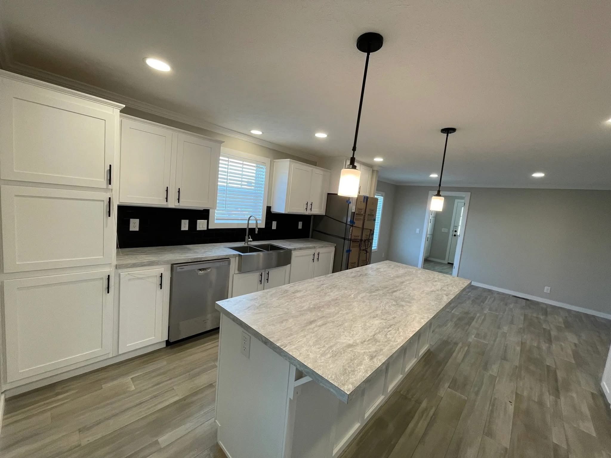 Spacious modern kitchen with white cabinets and gray countertops. Features a large central island, pendant lighting, stainless steel appliances, and wooden flooring.