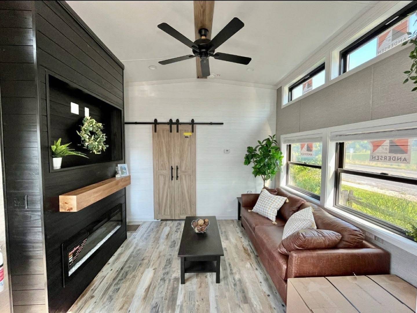 Cozy living room with a brown leather sofa, rustic wood coffee table, and large windows. Black fireplace, ceiling fan, and wooden sliding door enhance the modern vibe.
