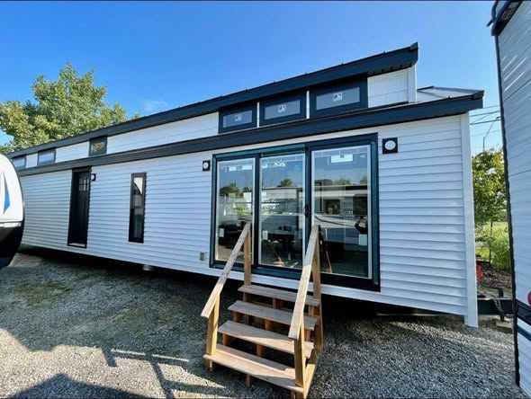 Modern tiny home with white siding, large glass doors, and a wooden staircase. Set in a sunny outdoor area, it conveys simplicity and minimalism.